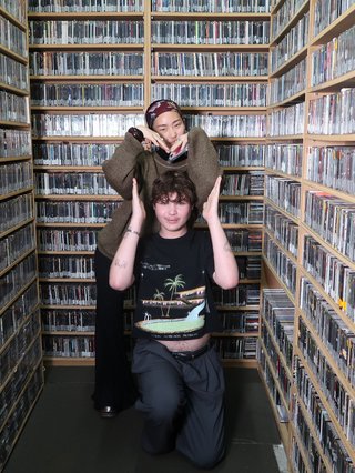 Ethan and Yvonne in the music library at the studio. In the background, you can see shelves of CDs lining the walls. Ethan is crouched down on the floor and Yvonne is standing behind him - they are making a heart with their hands.