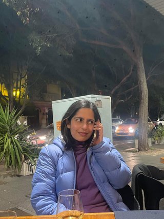 An image of a girl on the phone looking to her right and smiling. She has medium length dark hair and is wearing a light blue puffer jacket, sitting at a table on the street with a glass in front of her.