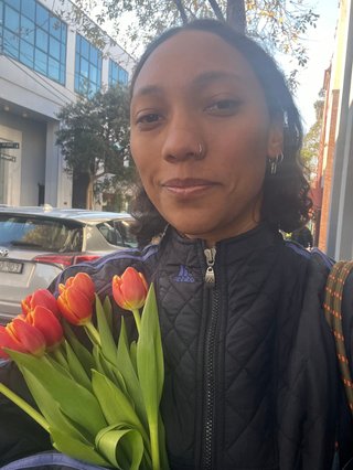 Bri is wearing a black puffer jacket and holding a bouquet of pink and yellow flowers.