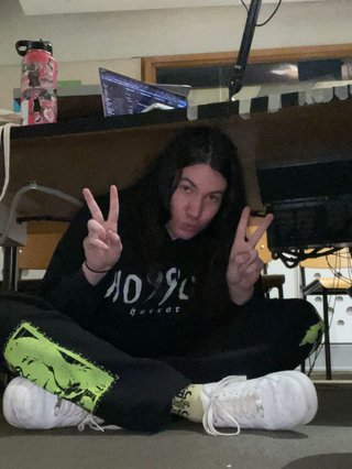 Jim sitting cross-legged under the desk in the fbi studio. Because they are a large human they are very hunched over, but still pouting and throwing up a peace sign to look cute for the camera.