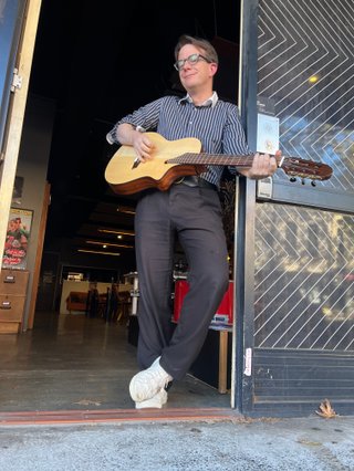 Geoffrey O'Connor standing in the doorway to fbi radio, strumming his guitar