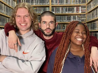 Benny, Guano C, and Ify stand in the fbi.radio music library. All three are looking at the camera, and Ify and Benny are smiling. Guano C looks fake confused. He stands in the middle with his arms over Ify and Benny's shoulders.