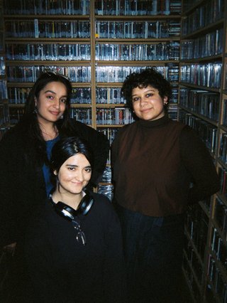 A film photo of Inez, Sehej and Shareeka in the music library. The lighting is off, only illuminated by the flash. Theya re all slightly smiling and lookign directly into the lens.