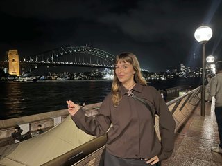 Izzy Page stands above looking down at Opera Bar. She poses in front of the Harbour Bridge in the far distance, pointing two fingers at it