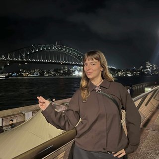 Izzy Page stands above looking down at Opera Bar. She poses in front of the Harbour Bridge in the far distance, pointing two fingers at it