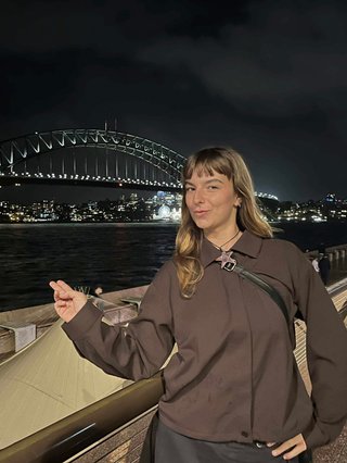 Izzy Page stands above looking down at Opera Bar. She poses in front of the Harbour Bridge in the far distance, pointing two fingers at it