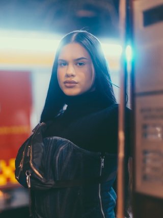 Jubah stands demure and strong in front of the camera. She's wearing a black patent corset on top of a black top. Her hair is slick and long, face card fierce