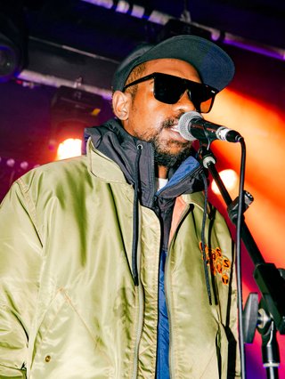 A portrait shot of Keanu Nelson singing into a microphone, shot from the waist up. He is wearing black sunglasses and a green jacket over a hoodie.