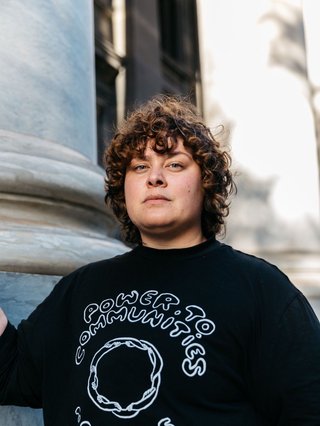 Latoya Aroha Rule stands firm in front of a colonial building looking directly at the camera. They wear a black sweater with white hand drawn font that says "Power to Community"