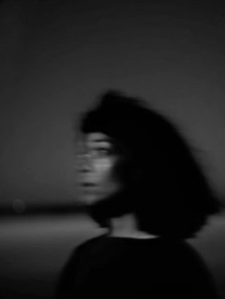 A black and white blurry portrait of Leah Avene. They are side, looking into the night sky with the ocean waves in the background