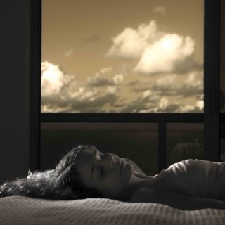 A side profile of Erika de casier lying down in bed, from the torso up. She is in black and white, and behind her an open window shows clouds in sepia colour