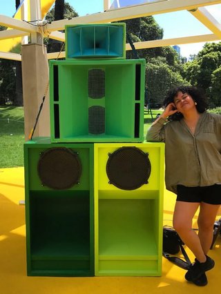 Lulu leans on her hand built sonic sound system. The amplifiers are different shades of green and tower over here. She is wearing green and black and smiling