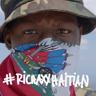 The album cover of Mach-Hommy's album #RICHAXXHAITIAN. It is a close up photo of Mach-Hommy wearing a bandana over the bottom half of his dface and a camo print bucket hat on his head. His facial expression seems neutral.