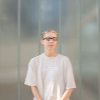 Mara stands in front of a frosted glass wall wearing a cream coloured t-shirt and black glasses. She is looking past the camera, upwards to the right.