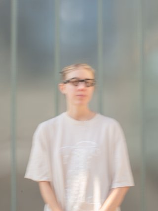 Mara stands in front of a frosted glass wall wearing a cream coloured t-shirt and black glasses. She is looking past the camera, upwards to the right.