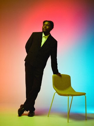 Musonga Mbogo, a black man with locs, leans against a chair and is lit my multicoloured light.