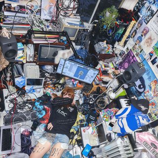 Ninajirachi covering her face, shot from above. she is in a croweded room packed full of laptops, wires and other electronics
