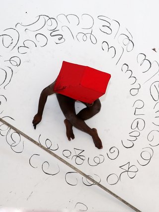 A still of an endurance performance by Dalit artist Sajan Mani. A dark-skinned body we see top-down sits cross-legged. Charcoal writing around him, a red hat and a large stick across the floor
