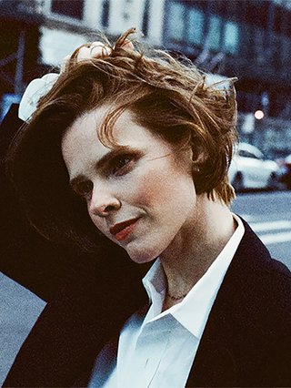 Cassandra Jenkins stands in a city street, they have auburn hair with their right hand bunching some up, they look down and off camera. They are wearing a black suit Jacket and a white collared shirt.