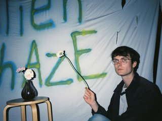 Other Joe sits to the right of frame, eyes acast, holding a flower with a long stem. He is sitting in front of a white sheet with letters painted in green and blue paint. A vase sits on a small stool to the left of frame.