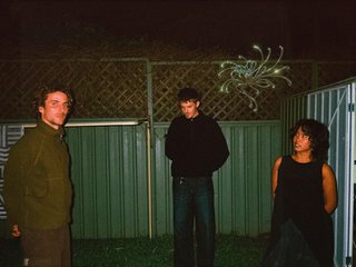 A greeny-grainy film photo of Bela, John and Bindi at night. They are standing in a backyard on grass, in front of a green fence