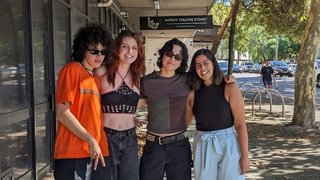The three members of MUNA stand on the street outside fbi.radio with Tanya Ali. All four of them are facing the camera and smiling in dappled sunlight, with arms around each other's backs.