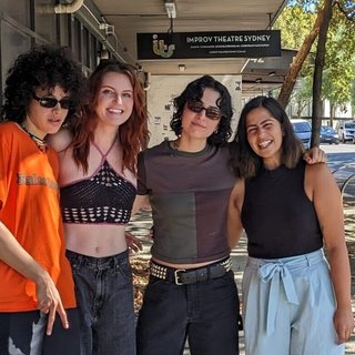 The three members of MUNA stand on the street outside fbi.radio with Tanya Ali. All four of them are facing the camera and smiling in dappled sunlight, with arms around each other's backs.