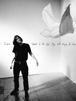 Album cover of Porridge Radio's album 'Clouds In The Sky They Will Always Be There For Me'. Black and white photograph of a person dressed in black standing alone in an empty room. They have a blurred face and slightly raised arms. A sculpture resembling a large bird made of wire and fabric hangs in the upper right quadrant.