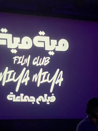 A still of an opening title at a Miya Miya screenign where Karim's silhouette stands in front of it. There is Arabic and English text artfully outlined on the screen.