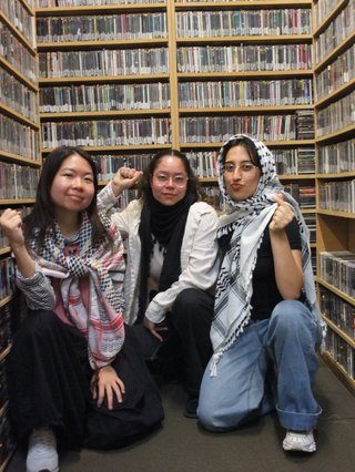 Joannie, Rand and Toobs squat in the fbi music library looking head on to the camera. Joannie has her wrist raised in solidarity, all are wearing Palestinian Keffiyahs