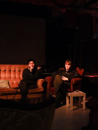 A photo of Shareeka and Ethan speaking live at The Red Rattler, a repurposed warehouse venue. They are on ragged chaise lounges and the lighting is red, and they're in front of an audience