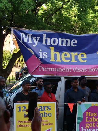 Protestor at the Refugee Encampment in Punchbowl. There are placards that read "My home is here, permanent visas now"