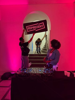 A candid snap from a Reprezent Radio fundraiser. The foreground features DJ decks, artists and the background sees two people afffixing a Reprezent sign to a door