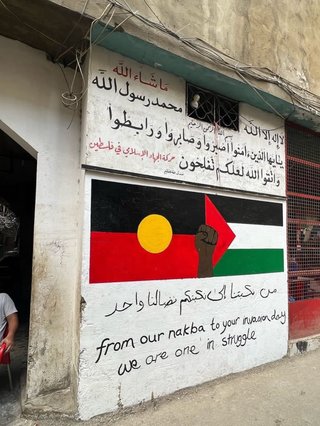 A photo of a mural in Jabilla Refugee camp home to many Palestinian refugees. There is a First Nations flag entwined with a palestinian flag. In both English and Arabic it reads "from our nakba to your invasion day we are one struggle"