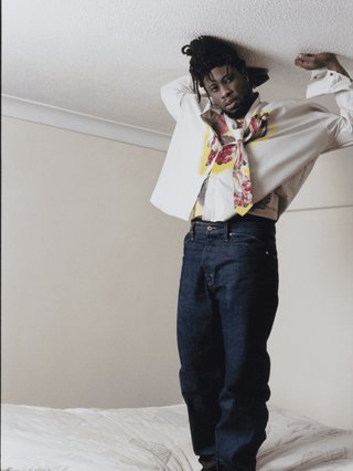 OB standing on a mattress with his hands above his head touching the roof. He is wearing a white shirt, jeans and neck scarf.