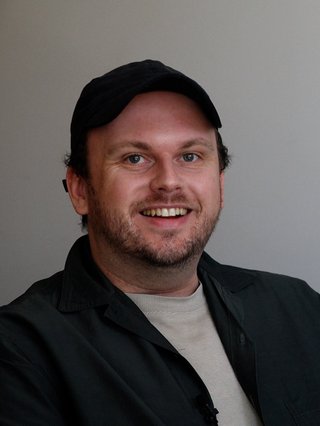 A portait shot of Samuel Van Grinsven. He is smiling at the camera, wearing a black cap, wearing a gray t shirt and black overshirt.