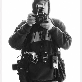 Sam 'Samoh' Stephensen, in Black and White, taking a photo, with photography equipment around their waist.