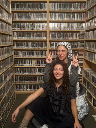 Sara and Malaak squatting on ground in fbi music library. Sara is making peace signs behind Malaak, they are both smiling.