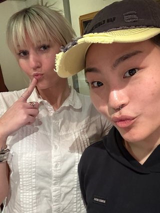 Two girls take a selfie. Girl on the right wears a baseball cap and wearing a black hoodie. Girl on the left is wearing a white button up shirt, and has her blonde hair tied up into a pony tail. She points her index finger to her lip.