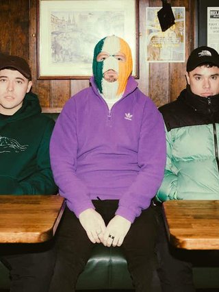 The band Kneecap sit against a wood-grained wall. The middle member, DJ Provai, is wearing a full-face balaclava adorned with colours of the Irish flag.