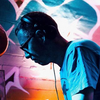 Simon Caldwell stands side on, looking downwards. He has headphones on. Behind him is a brightly lit and colourful brick wall with graffiti.