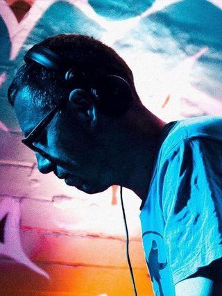 Simon Caldwell stands side on, looking downwards. He has headphones on. Behind him is a brightly lit and colourful brick wall with graffiti.