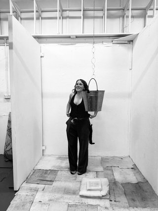 Black and white photo of Sophie standing in a gallery space. A bucket is hanging by a chain and is suspending in the middle of the picture. The floor is lots of overlaying fabric.