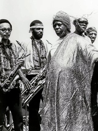 Black and white image of Sun Ra on his side, wearing a glittery robe and head dress looking forward, before four members of his Arkestra (band) hlding brass instruments.