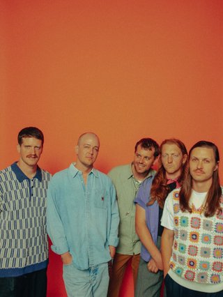 All five members of Surprise Chef stand side by side looking into the camera, before an orange-rust coloured backdrop