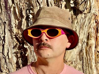 Tyson stands in front of a paperbark tree wearing orange sunglasses, a suede bucket hat and a pink t-shirt