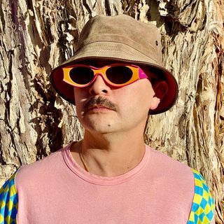 Tyson stands in front of a paperbark tree wearing orange sunglasses, a suede bucket hat and a pink t-shirt