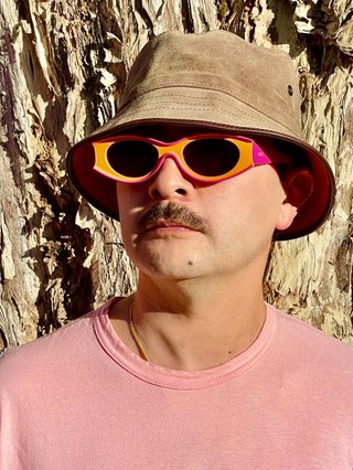 Tyson stands in front of a paperbark tree wearing orange sunglasses, a suede bucket hat and a pink t-shirt