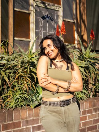 Tanya is standing in front of plants and a brick fence on a sunny day. She is looking to the left and smiling