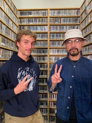 Béla and Troy stand next to each other in the FBi music room, both holding up peace signs with their hands. On the left, Béla is wearing a navy hoodie. On the right, Troy is wearing a dark denim shirt, and a white hat. Behind them and surrounding them are shelves of CDs.